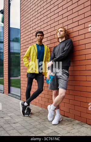 Zwei junge Männer in stylischer Sportkleidung posieren selbstbewusst vor einer eleganten Ziegelwand. Stockfoto