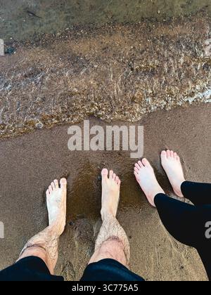 Mann und Frau stehen barfuß auf Sand, während sich die Welle nähert. Stockfoto