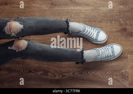 Frauenbeine tragen zerrissene Jeans und Turnschuhe, die auf einem Holzboden sitzen. Draufsicht Stockfoto