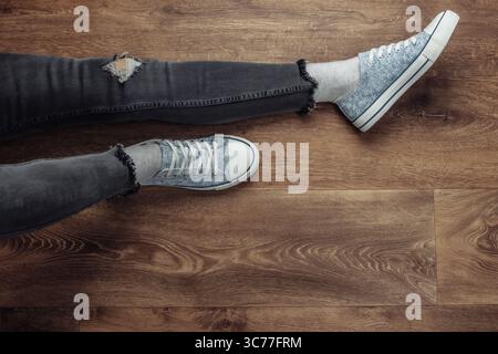 Frauenbeine tragen zerrissene Jeans und Turnschuhe, die auf einem Holzboden sitzen. Draufsicht Stockfoto