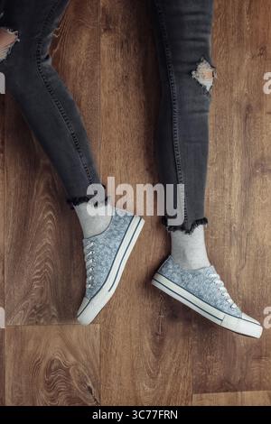 Frauenbeine tragen zerrissene Jeans und Turnschuhe, die auf einem Holzboden sitzen. Draufsicht Stockfoto