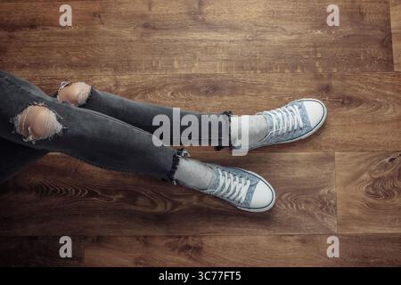 Frauenbeine tragen zerrissene Jeans und Turnschuhe, die auf einem Holzboden sitzen. Draufsicht Stockfoto