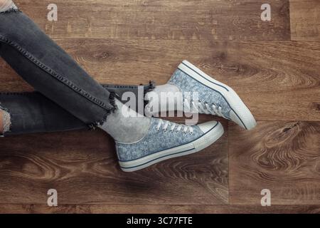 Frauenbeine tragen zerrissene Jeans und Turnschuhe, die auf einem Holzboden sitzen. Draufsicht Stockfoto