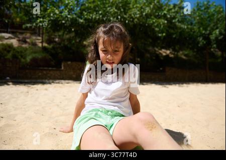 Lächelndes Mädchen in einem weißen Oberteil und grünen Shorts auf einer sandigen Oberfläche, umgeben von üppigen Bäumen und sonniger Umgebung in einem Park. Stockfoto
