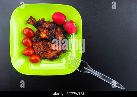 Grüner Plastikteller mit einem Stück gegrilltem saftigem Fleisch mit Gemüse auf schwarzem Hintergrund. Frittiertes Fleisch mit Tomaten und Radieschen. Hochwertiges Foto Stockfoto