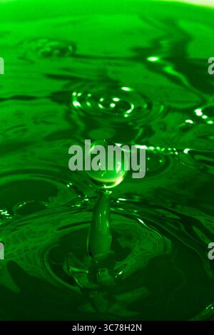 Ein einziger Wassertropfen fällt in eine leuchtend grüne Flüssigkeit, erzeugt einen schönen Spritzer und kräuselt sich über die Oberfläche, die in einer detaillierten Nahaufnahme erfasst wird. Stockfoto