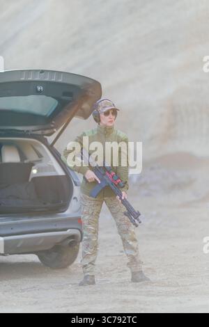 Soldatinnen stehen neben dem Fahrzeug mit offenem Kofferraum, halten Gewehr, tragen Tarnuniform, Gehörschutz und Sonnenbrille in Wüstenumgebung Stockfoto