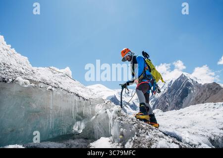 Bergsteiger in pulsierenden Gewerken überqueren Gletscherspalten während des Lenin Gipfels unter klarem Himmel mit schneebedeckten Gipfeln im Hintergrund. Extreme Akti Stockfoto