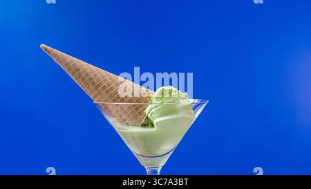 Eine erfrischende Portion Matcha-grüner Eiscreme wird elegant in eine Waffelhülle gelegt, die am Rand eines Glases ausbalanciert ist. Der leuchtend blaue Hintergrund wird noch verbessert Stockfoto