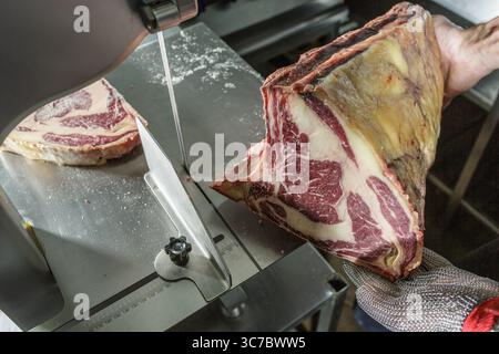 Rohes, trockengereiftes Ribeye-Steak, das vom Metzger mit Schutzhandschuh auf Stahltisch mit einer Metallsäge geschnitten wird Stockfoto