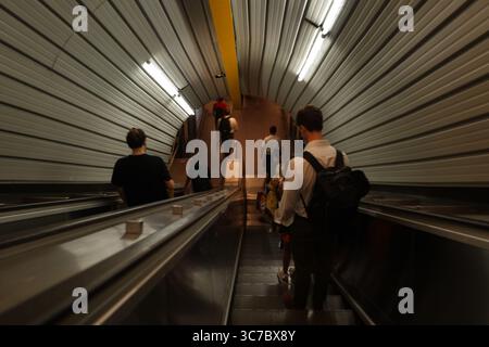 Passagiere steigen auf Rolltreppen in einer modernen U-Bahnstation in Rom ab. Der metallische, gerippte Tunnel schafft eine futuristische Kulisse für die bewegte Krähe Stockfoto