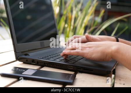 Hände, die auf einer Laptop-Tastatur auf einem Holztisch im Freien tippen, mit einem Smartphone daneben, was auf Fernarbeit oder Freiberufung hindeutet Stockfoto
