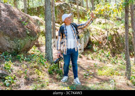 Der Fotograf steht mitten in einem üppigen Kiefernwald und lächelt während eines Videogesprächs. Mit seiner Kamera in der Hand bringt er Technologie und Natur in Einklang Stockfoto