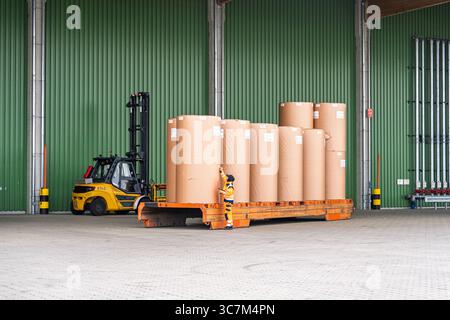 Arbeiter, der große Papierrollen auf einem Flachbettanhänger in einer Lagerumgebung sichert, Industrie Stockfoto