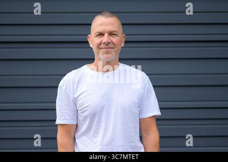 Porträt eines selbstbewussten älteren Mannes mit kurzen grauen Haaren und Bart, der vor einer modernen dunkelgrauen Wand steht. Stockfoto