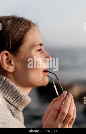 Frau Brillenprofil, nachdenkliche junge Frau, die draußen am Meer eine Brille hält. Stockfoto