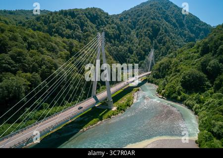 Eine Kabelbrücke überspannt einen Fluss, der von üppigen grünen Bergen umgeben ist, mit Autos, die entlang der Straße fahren Stockfoto