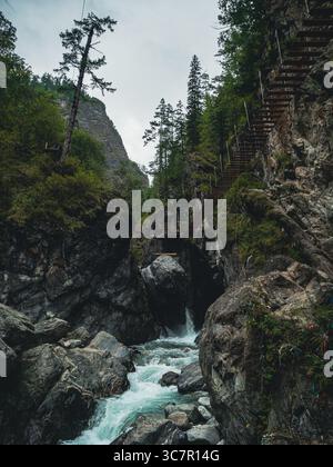 Wasser rauscht über glatte Steine am Fuße der zerklüfteten Klippen, während sich eine Holzbrücke über die felsige Landschaft erstreckt. Stockfoto