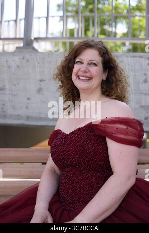 Frau in pompösem rotem Abendkleid sitzt auf der Bank und lächelt Stockfoto