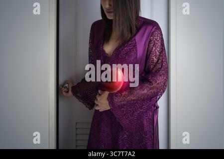 Eine Frau in einem lila Nachthemd aus Spitze, die an Schlaflosigkeit durch Verstopfung leidet, wird gesehen, wie sie ihren Bauch in den Griff nimmt, nachdem sie aus dem Badezimmer kam Stockfoto