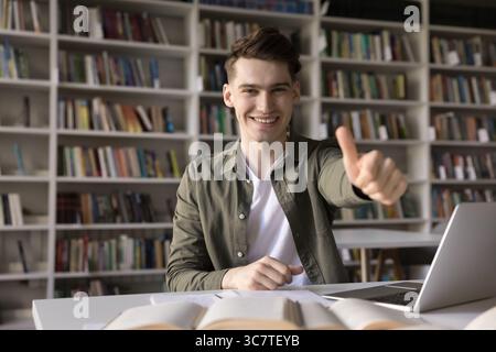 Ein lächelnder männlicher Schüler sitzt am Schreibtisch des Klassenzimmers und zeigt die Daumen hoch Stockfoto