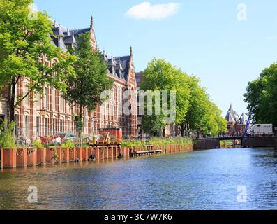 Hübsche Aussicht auf einen Kanal in Amsterdam, niederlande, mit einem Kolonialgebäude aus rotem Backstein und einer Bogenbrücke in der Ferne Stockfoto