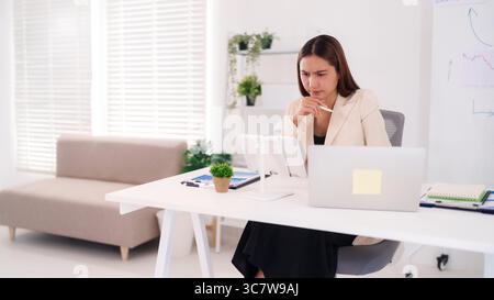 Durchdachte asiatische Erwachsene Frau mit Schwerpunkt auf Marktdaten und Geschäftsstrategie mit Tablet und Laptop in einer hellen, modernen Büroumgebung Stockfoto