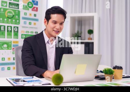 Professioneller, erwachsener asiatischer Mann, der auf einem Laptop lächelt und aktiv an seinem modernen Bürotisch für Nachhaltigkeit und umweltfreundliche Initiativen engagiert ist Stockfoto