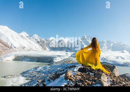 Eine junge kaukasische Frau, von hinten gesehen, in einem leuchtend gelben Kleid sitzt auf einem felsigen Bergrücken mit Blick auf einen Gletscher und schneebedeckte Berge Stockfoto