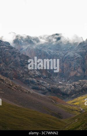 Eine vertikale Aufnahme eines zerklüfteten, felsigen Bergtals mit steilen Hängen und Wolken, die sich an den Gipfeln schmiegen, mit einem kleinen Bach, der unten fließt Stockfoto