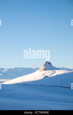 Eine Straße schlängelt sich durch eine riesige schneebedeckte Landschaft mit einem markanten felsigen Berg im Hintergrund, der von der Morgensonne beleuchtet wird Stockfoto