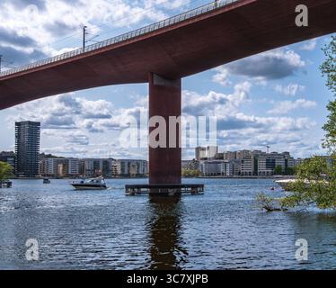 Stockholm, Schweden - 5. Juni 2025: Malerischer Blick auf eine moderne Brücke, die über einen ruhigen Fluss überspannt, mit Booten darunter. Der Himmel ist teilweise bewölkt, Stockfoto