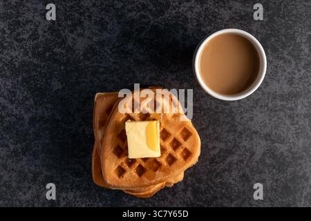 Blick von oben auf köstliche herzförmige Waffeln, gekrönt mit einem Würfel schmelzender Butter, serviert zusammen mit einer Tasse Kaffee, alles auf einer dunklen, strukturierten Oberfläche Stockfoto