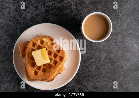 Direkt über dem Kopf mit Blick auf einen Stapel goldener herzförmiger Waffeln mit einem schmelzenden Stück Butter auf einem gesprenkelten Teller, der im Dunkeln steht Stockfoto