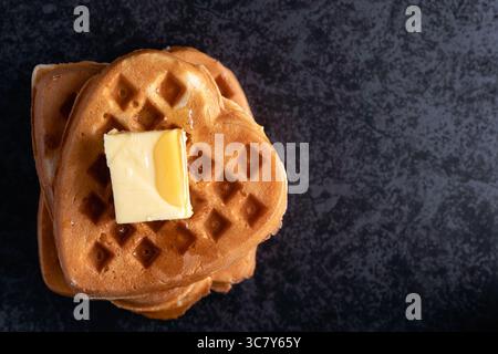 Hochwinkel-Nahaufnahme eines Stapels herzförmiger Waffeln, die großzügig mit einem Quadrat schmelzender Butter und einem Tropfen Sirup auf dunklem Hintergrund belegt sind Stockfoto