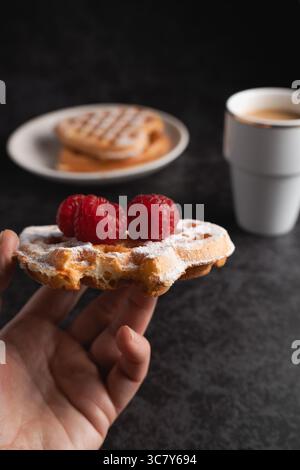 Nahaufnahme einer Hand mit einer einzigen goldenen Waffel, verziert mit Puderzucker und zwei Reifen Himbeeren, mit verschwommenem Hintergrund Stockfoto