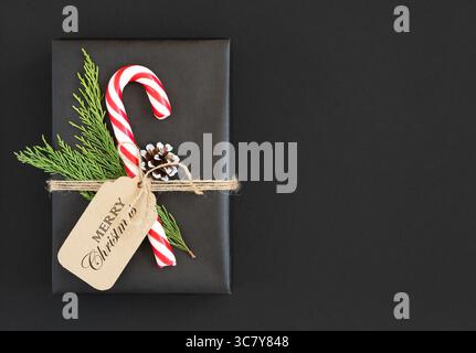 Schwarze Geschenkbox mit Zuckerrohr, Zedernzweig, Kegel und Etikett mit dem Text Merry christmas auf dunklem Hintergrund. Platz für den Text. Stockfoto