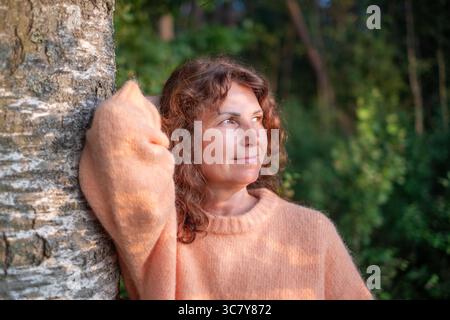 Die Frau mit lockigen Haaren in einem weichen Pullover lehnt sich an einen Baum, in die Natur eingetaucht, umgeben von Grün, und fängt das Wesen ein Stockfoto