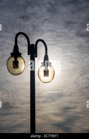 Eine Straßenlaterne mit zwei Glühlampen, von denen eine durch die Sonne direkt dahinter beleuchtet erscheint und einen auffälligen visuellen Effekt erzeugt. Stockfoto