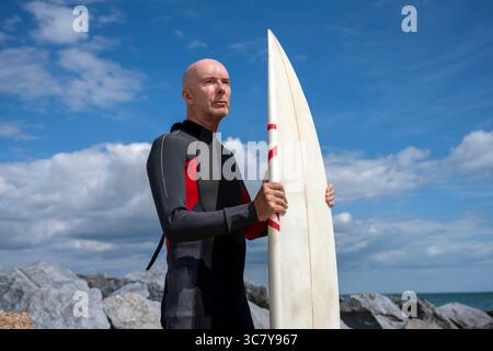 Surfer, der am Strand steht und ein Surfbrett in einem Neoprenanzug hält Stockfoto