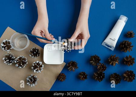 Person, die Tannenzapfen Weihnachten auf blauem Hintergrund malte. Vorbereitung der Weihnachtsdekoration. Stockfoto