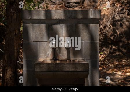 Alter Trinkwasserbrunnen aus Stein mit zwei Metallhähnen, im Freien zwischen Bäumen gelegen, in natürlichem Sonnenlicht mit Schatten, reflektiert eine rustikale und utilita Stockfoto