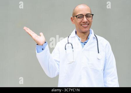 Porträt eines Reifen männlichen Glatzenarztes mit Labormantel und Brille mit Stethoskop um den Hals, posierend vor neutralem Wandhintergrund. Ideal für ihn Stockfoto