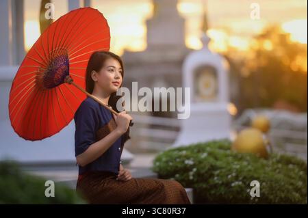 Seitenansicht einer jungen Thai-Frau, die an einer Wand neben einem Tempel sitzt, der einen Sonnenschirm hält, Thailand Stockfoto