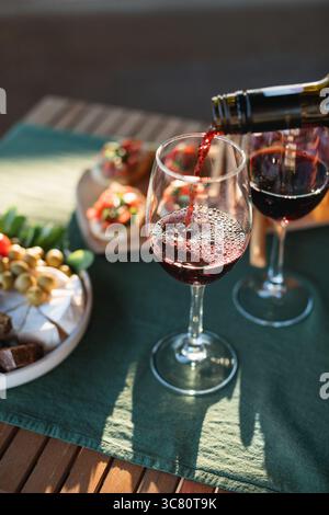 Rotwein, der auf einem Gartentisch in ein Glas gegossen wird, mit verschiedenen Aperitivo-Snacks im Sommer Stockfoto