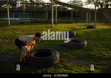 Mann mit schwarzen Shorts, der auf dem Feld hockt, der Traktorreifen in der Nähe des Hindernisrahmens umkippt, Kopierraum Stockfoto
