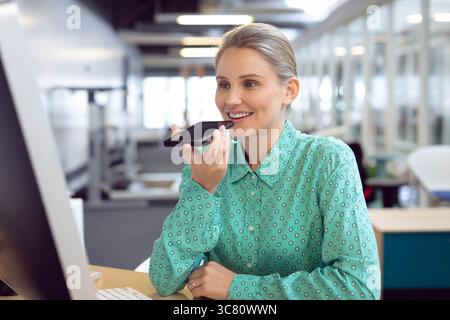 Smartphone, das Sprachnachrichten in der Nähe des Computermonitors und der Tastatur am offenen Bürotisch sendet Stockfoto