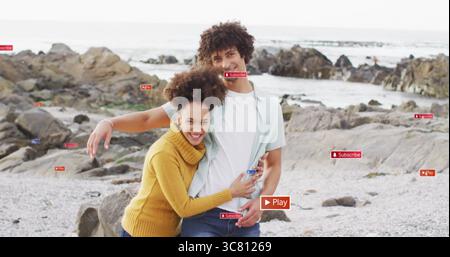 Lächelndes Paar umarmt sich an der felsigen Küste in Jeans und gelbem Pullover, mit Kieseln und Meer Stockfoto