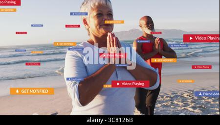 Yoga-Partner in sportlicher Kleidung, die Gebetsposen am Meeresrand mit Social-Media-Alerts durchführen Stockfoto