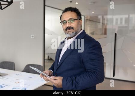 Ernsthafter nachdenklicher reifer Geschäftsmann, der im Sitzungssaal des Büros steht und ein Tablet hält Stockfoto